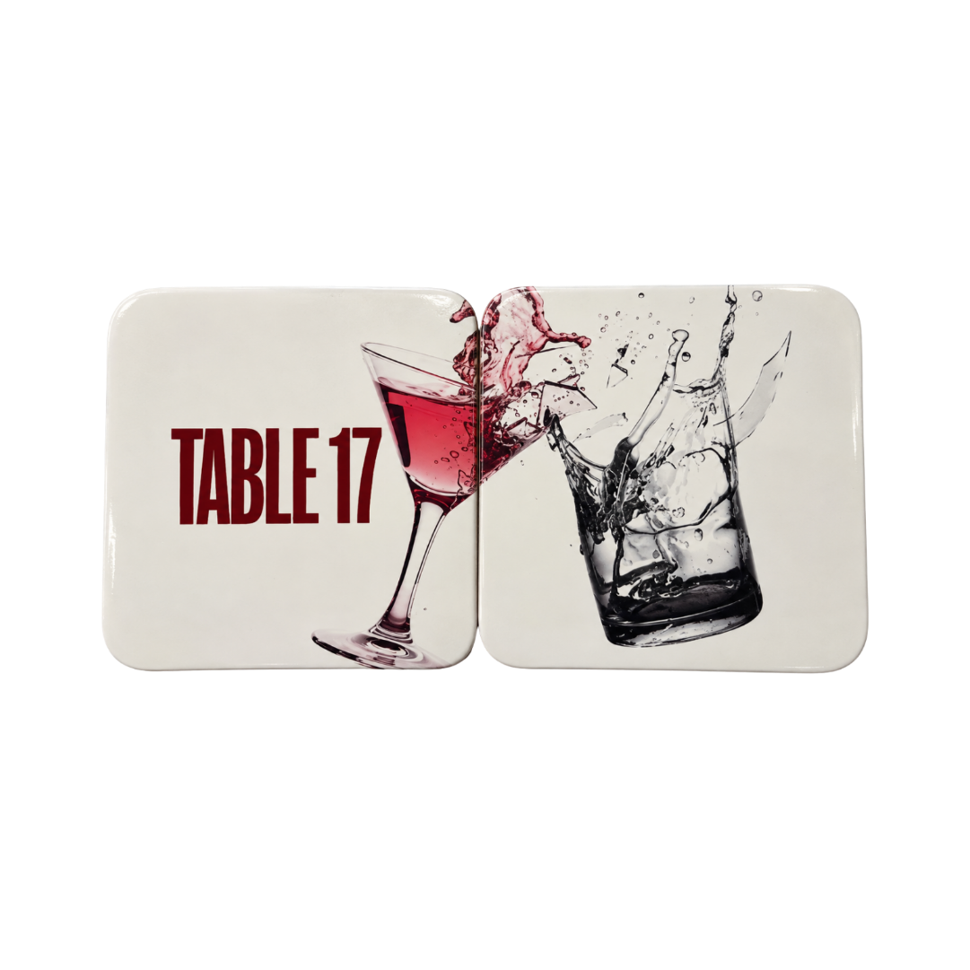 A two-piece table coaster set with rounded corners. The left coaster is off-white with the text 'TABLE 17' in dark red and an illustration of a martini glass with red liquid splashing.