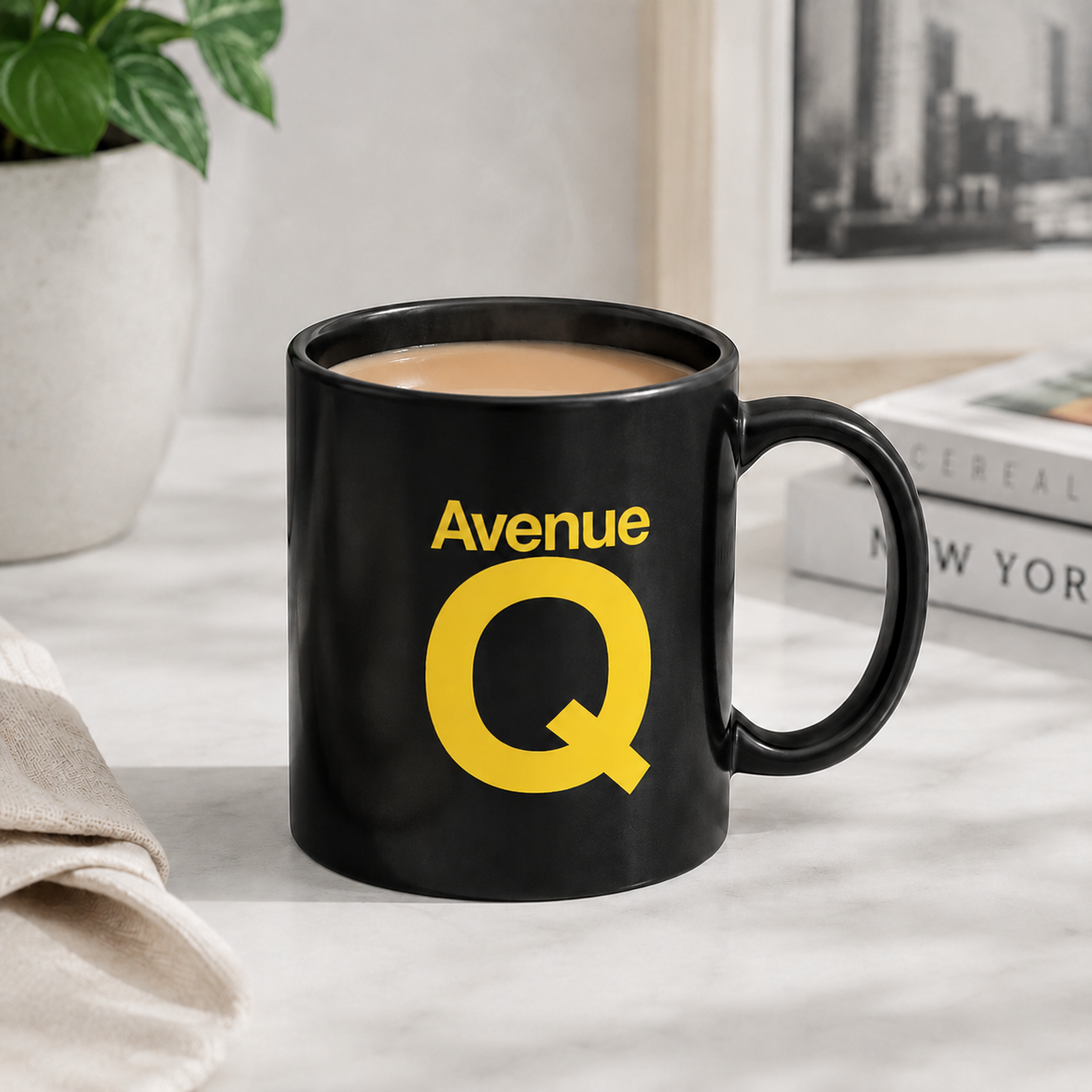 A black ceramic coffee mug with a yellow 'Avenue Q' graphic, filled with coffee and set on a marble countertop in a kitchen setting.